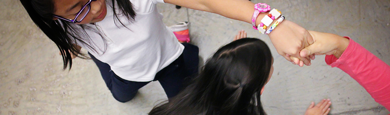 Three girls play a game. Two of the girls are holding hands up high like a bridge and the third girl is about to pass under them.