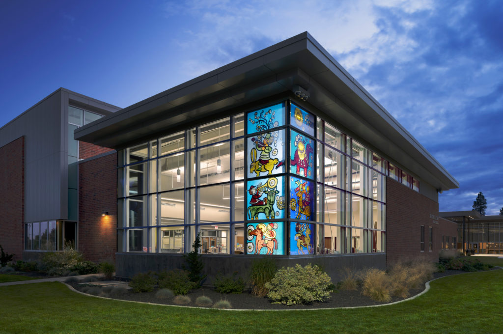 Eight stained glass windows form the corner of double-height windows in a school library, shown from the outside at dusk. The windows feature a series of people playing musical instruments and riding animals.