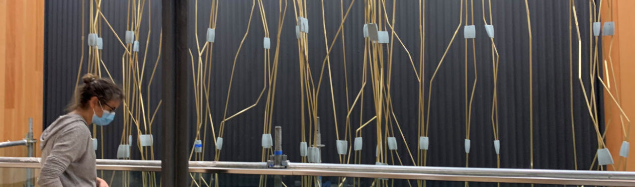 A woman is installing an artwork made of several bunches of gold-colored cables hanging in front of a dark grey wall.