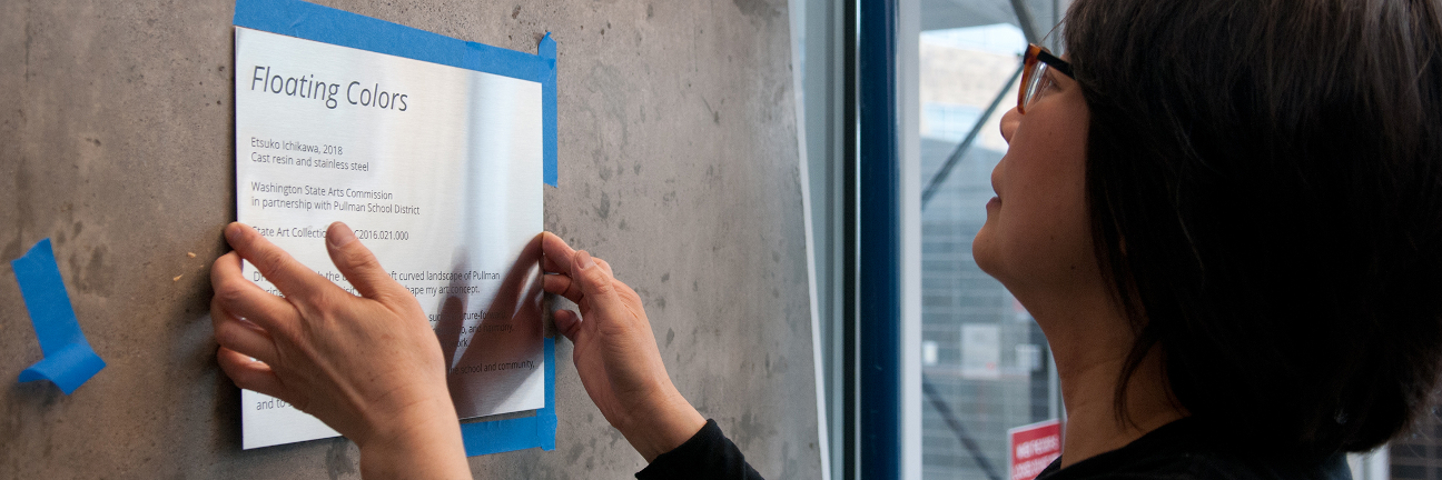 A woman attaches a metal plaque for an artwork onto a concrete wall.