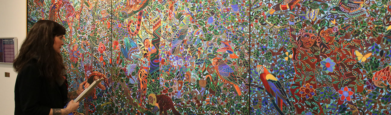A woman looks at a a large, wide painting of a detailed jungle scene and makes notes.