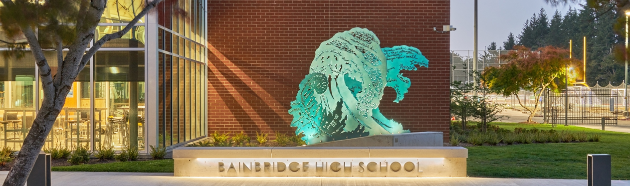 A large fern frond and two large waves made of flat, aluminum in shades of blue stand parallel to each other on a concrete base in front of a brick school building. Each element also has words and patterns cut into it.