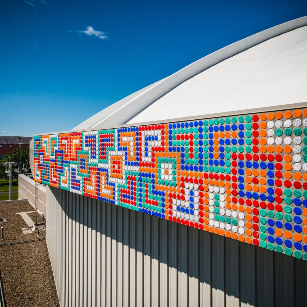 Partial view of a band of geometric patterns made of thousands of 3-inch industrial highway reflectors encircling the circular roof of a large concrete building. There is a very blue sky behind the SunDome building.