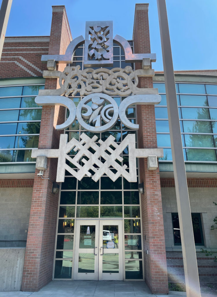 "A Collection" stainless steel, aluminum, and concrete sculpture by Harold Balazs. A large sculpture is integrated into a two-story brick building, above the entry doors. The sculpture features four rows of abstract patterns on the building façade.