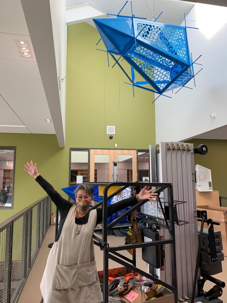 The artist is standing with her large, blue, metal, crystal-shaped sculpture, that is hanging above her in a large light well, during the artwork installation. Her arms are raised up and out in a triumphant and happy pose. There is installation equipment around her as well.