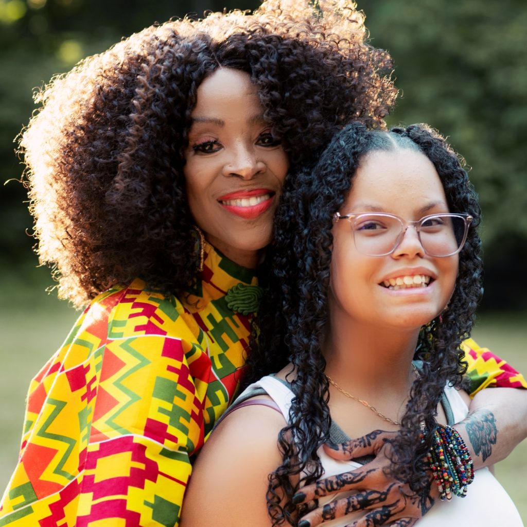 Franchesska Berry is a master of West African and African diaspora dance. and cultural herstory. Apprentice is Nola Taylor. In this photo, Franchesska stands behind Nola and gives her a gentle hug.