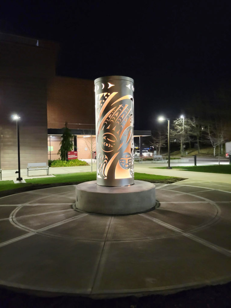 A sculpture installed on a college campus plaza is shown at night. The metal cylindrical sculpture has cut-out faces and patterns on it, in the Northwest Coast style. It also contains an orange cylinder within it that is lit up from within and glows. "ININYAX’A7N (Thunderbird)" by James Harry.